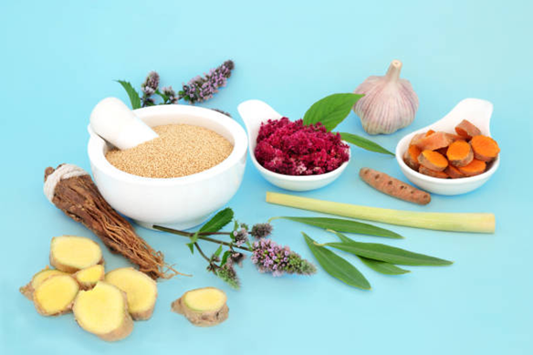 Assorted herbs and roots arranged on a blue surface, including ginger slices, turmeric, lemongrass, garlic, and bowls with ground powders and red crushed herb.