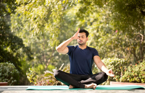 Man sits cross‑legged on a yoga mat outdoors, eyes closed, hand against his forehead in a meditative gesture.