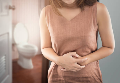 Person standing indoors, clutching their abdomen as if in stomach pain, with a bathroom visible in the background.