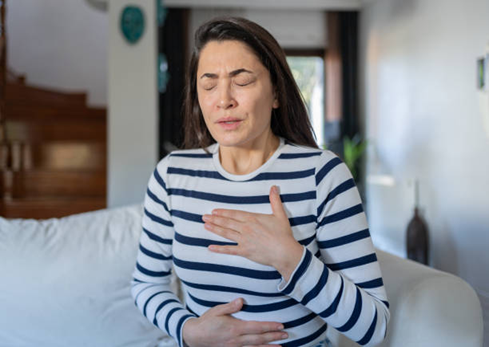 Woman sitting on a couch indoors with eyes closed, one hand on her chest and the other on her abdomen, appearing to breathe deeply or feel chest discomfort.