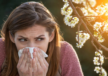 Woman outdoors blowing her nose into a tissue, with blooming branches and sunlight in the background, suggesting seasonal allergies.