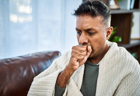 Man sitting on a sofa wrapped in a blanket, coughing into his fist with a concerned expression at home.