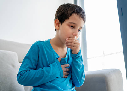 Boy sitting on a sofa, hand over mouth, looking worried or anxious in a blue shirt.