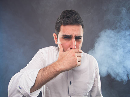 Man in a white shirt covering his mouth with his hand, appearing to cough, with blue smoke in the background.