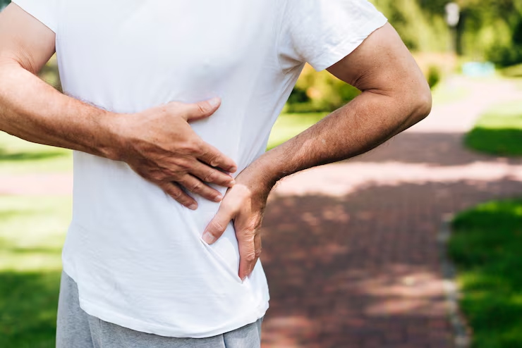 Person outdoors in a white T-shirt touching their side, suggesting abdominal discomfort or a side ache.