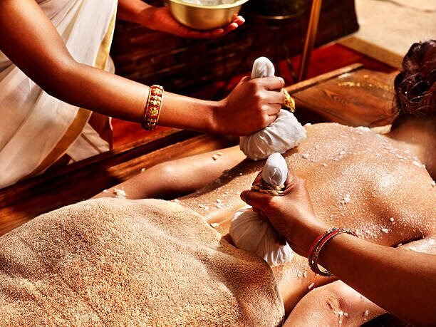 Two spa attendants scrub a back with cloth sachets as a person lies prone on a wooden massage table, covered in coarse scrub.”,