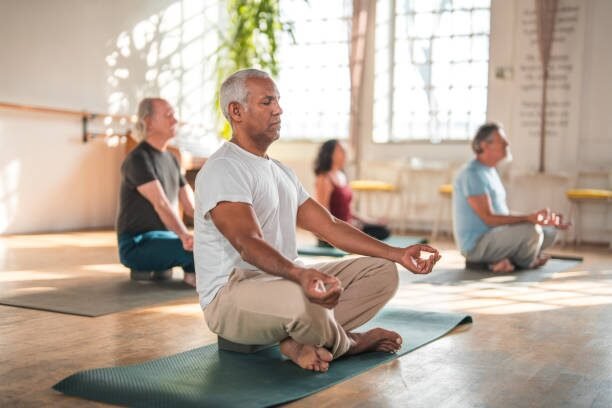 Group of adults meditating in a sunlit yoga studio, seated on mats in lotus/meditative pose with eyes closed and hands in mudra.