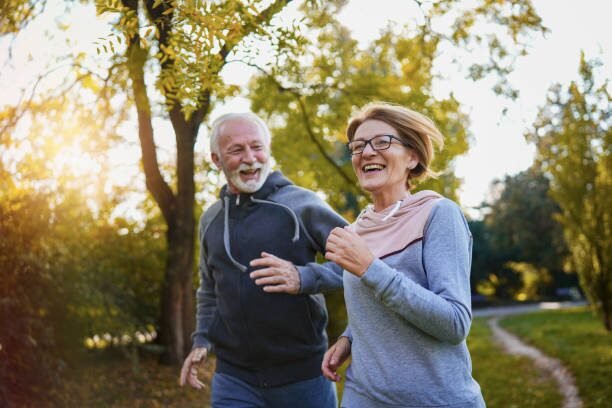 An older couple jogging together on a sunny park path, smiling and enjoying the walk.