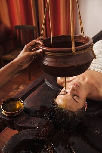 A therapist pours warm oil from a clay pot onto a woman's face during a spa facial.