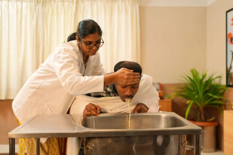 A female caregiver in a white coat tilts a man’s head over a sink, pouring liquid into his mouth.