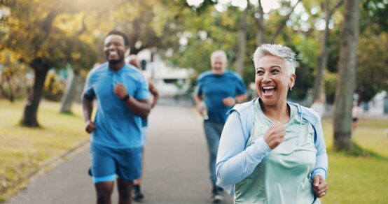 Smiling older woman running in a sunny park, with two men jogging behind her in the distance.
