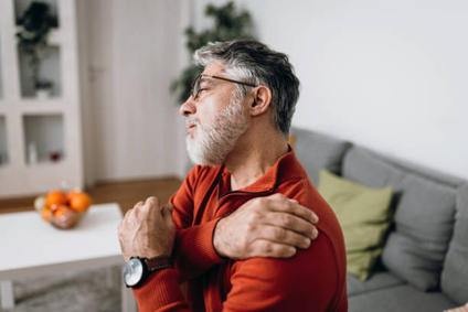 Older man with gray hair and beard in a red sweater stretches by crossing his arms over his chest in a living room setting.