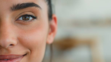 Close-up of a smiling woman's face, showing her left eye, eyebrow, and cheek with a blurred background behind her.