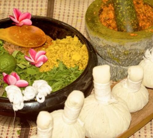 Herbal spa setup: a dark bowl filled with colorful ground spices and herbs, pink flower decorations, a wooden spoon resting on the rim, and white tied herb bundles on a wooden surface.
