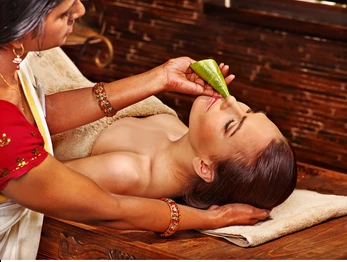 Therapist performing a facial treatment on a client lying on a spa bed; a green leaf rests on the client's lips.