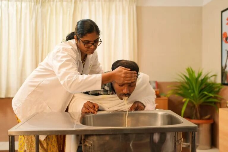 Female caregiver in a white coat rinses a man's head over a stainless-steel sink in a clinic-like room, assisting with washing.