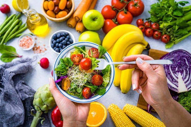 Person holding a bowl of fresh salad with cherry tomatoes, greens and sprouts, surrounded by colorful vegetables and fruit on a kitchen table