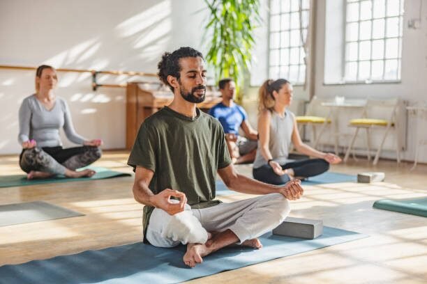 Group of adults meditating on yoga mats in a bright studio; front person sits cross‑legged with hands in a mudra.