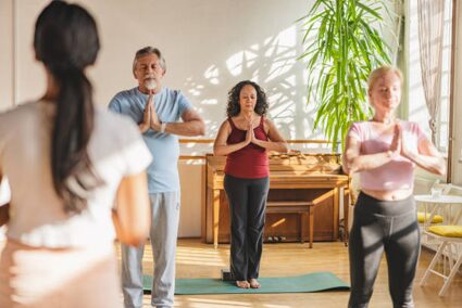 A small group of adults in a sunlit yoga studio, standing in prayer pose with hands together and a teacher at the back guiding them.