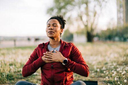 Person meditating outdoors with eyes closed, hands resting on chest in a park setting.