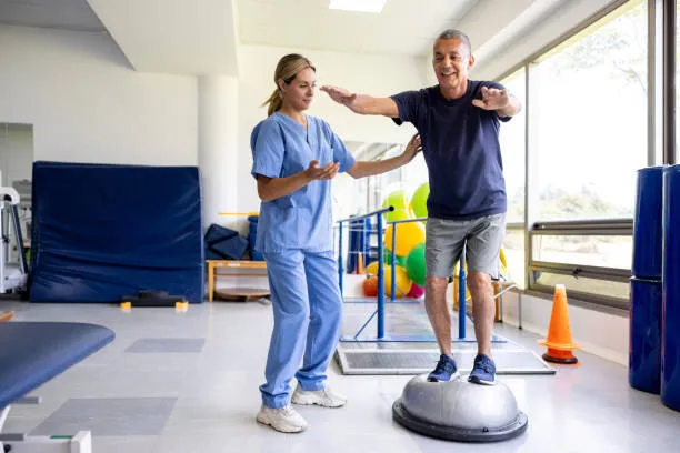 Therapist supervising an older man doing balance training on a wobble dome in a rehab gym.