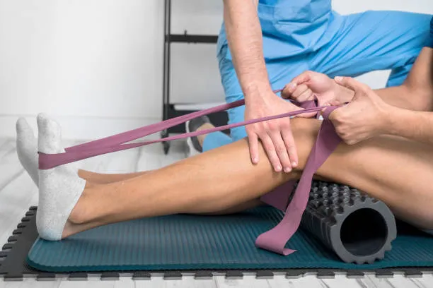 Physical therapist uses a purple resistance band to stretch a patient’s ankle while lying on a mat with a foam-roller nearby.