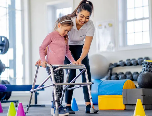 Young girl in a pink sweater uses a walker while a therapist guides her in a sunny therapy gym.