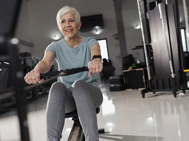 Older woman using a rowing machine in a bright gym, smiling at the camera.
