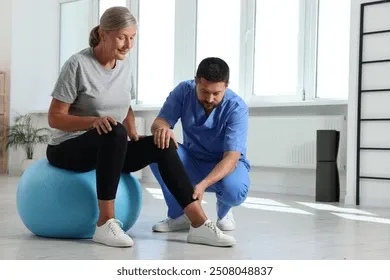 Older woman sits on a blue exercise ball, performing a knee bend while a trainer supports her in a bright studio environment.