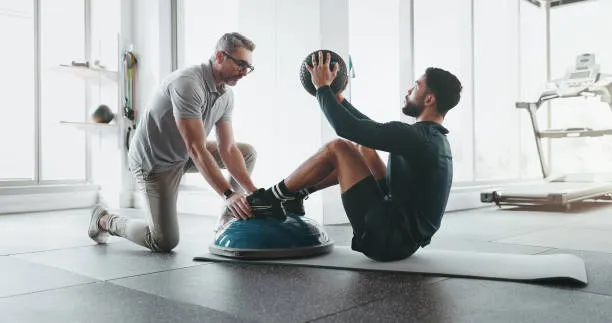 Fitness trainer kneels beside a client who is performing a core exercise on a BOSU balance ball in a bright gym there is a mat on the floor.