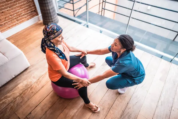 Therapist in blue scrubs supports a seated woman on a pink stability ball as they hold hands during a gentle exercise in a bright, airy room with a wooden floor and railing.