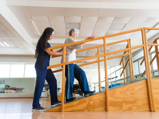A young woman helps an elderly man walk up a wooden ramp with orange handrails in a bright rehabilitation room.