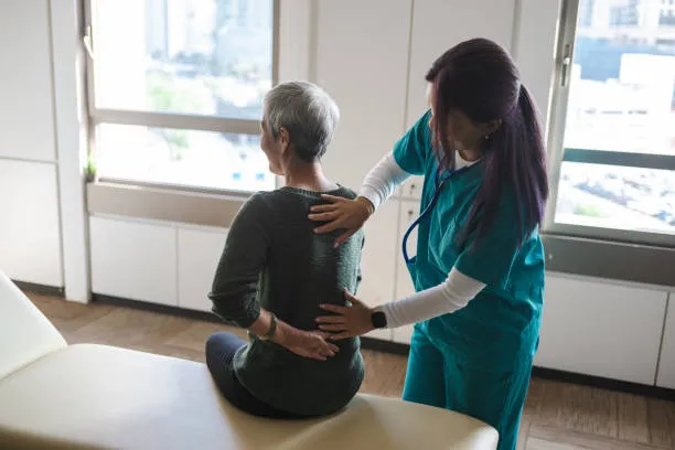 Female clinician in teal scrubs gently supports an elderly woman seated on an exam table during a back examination in a clinic.