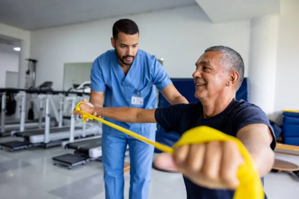 Physiotherapist in blue scrubs guides a man through a shoulder stretch with a yellow resistance band in a gym.