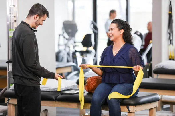 Therapist assists a smiling woman seated on a treatment table with yellow resistance bands in a clinic.