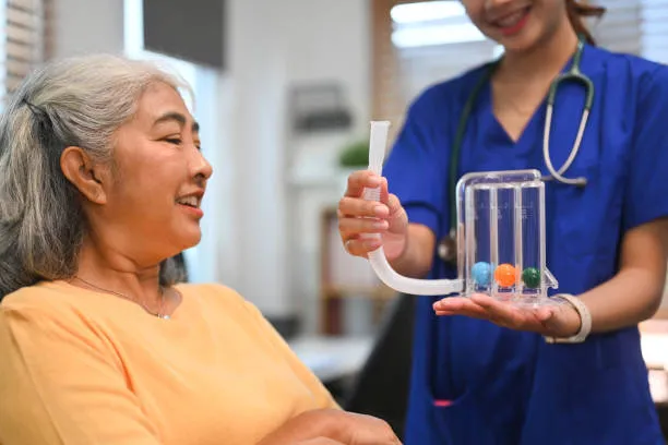 Nurse in blue scrubs and a stethoscope demonstrates a medical device with a curved tube and four colorful balls to an elderly smiling woman.