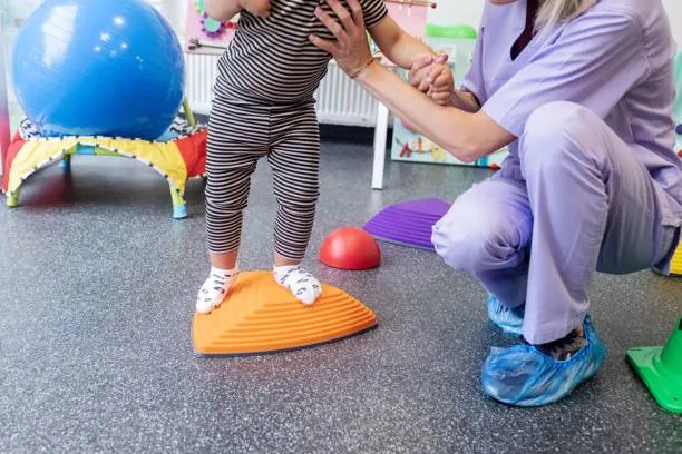 Child wearing stripes balances on an orange wobble dome while a caregiver supports them in a therapy room.