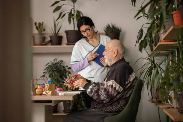 Caregiver assisting an elderly man with a hand-held toy at a table in a plant-filled room.