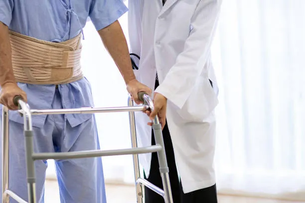 Patient wearing a lumbar back brace using a walker while a clinician assists nearby.