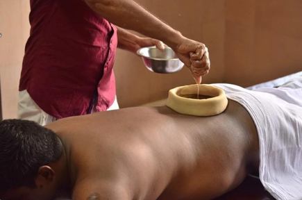Therapist pours oil onto a client's back during a massage while the client lies prone on a table, draped with a sheet.